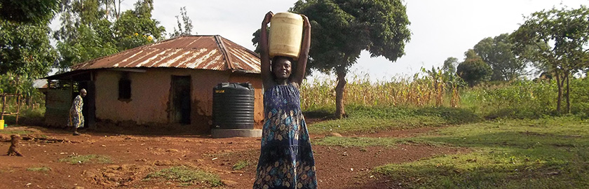 woman carrying a water bucket on her head and smiling at the camera. she is standing outside in front of a small structure in a field.